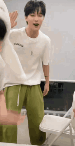 A Man In A White T-Shirt And Green Pants Is Standing Next To A Folding Chair . GIF