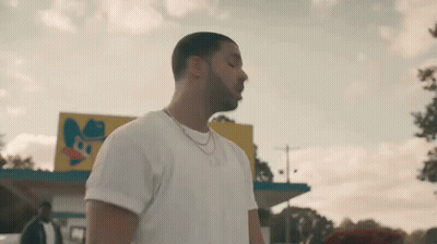 A Man In A White T-Shirt And Necklace Is Standing In Front Of A Fast Food Restaurant . GIF