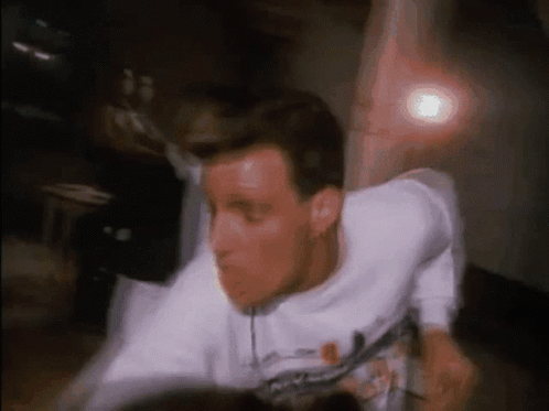 A Man In A White T-Shirt Is Dancing In Front Of A Light GIF