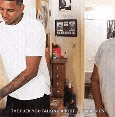 A Man In A White T-Shirt Is Standing In A Hallway Talking To Another Man . GIF