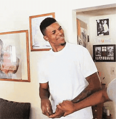 A Man In A White T-Shirt Is Standing In A Living Room Holding Another Man 'S Hand . GIF