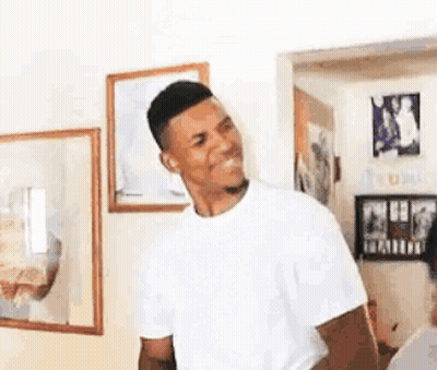 A Man In A White T-Shirt Is Standing In A Living Room With A Smile On His Face . GIF