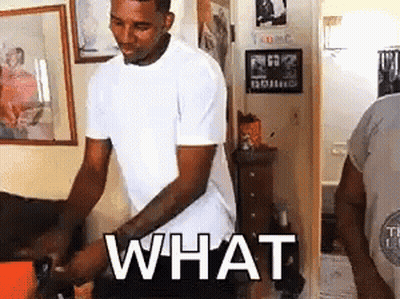 A Man In A White T-Shirt Is Standing In A Living Room With The Word What Written On His Chest . GIF