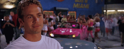 A Man In A White T-Shirt Is Standing In Front Of A Pink Car . GIF