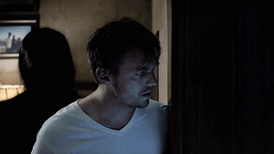 A Man In A White T-Shirt Stands In A Dark Room GIF