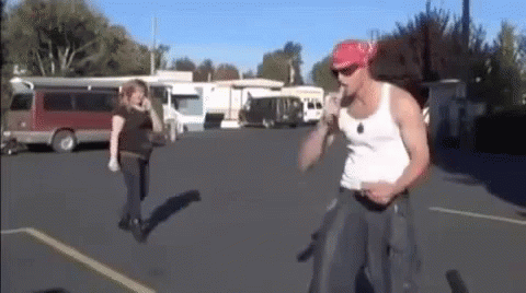 A Man In A White Tank Top And A Red Bandana Is Standing In A Parking Lot With A Woman Walking Behind Him . GIF