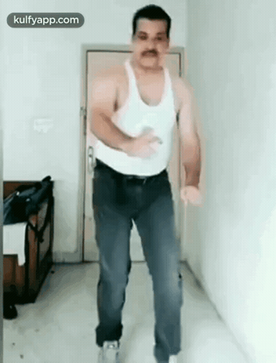A Man In A White Tank Top And Blue Jeans Is Dancing In A Room . GIF