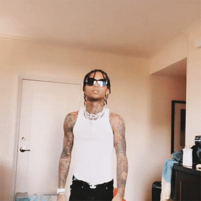 A Man In A White Tank Top And Sunglasses Stands In A Room GIF