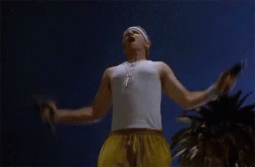 A Man In A White Tank Top And Yellow Shorts Is Standing With His Arms Outstretched In Front Of A Palm Tree . GIF