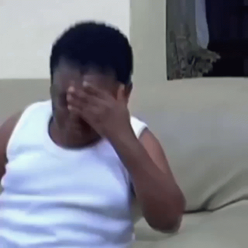 A Man In A White Tank Top Is Covering His Face With His Hands . GIF