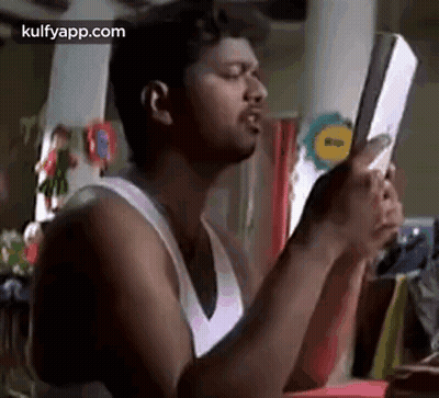 A Man In A White Tank Top Is Holding A Book In His Hands And Looking At It . GIF