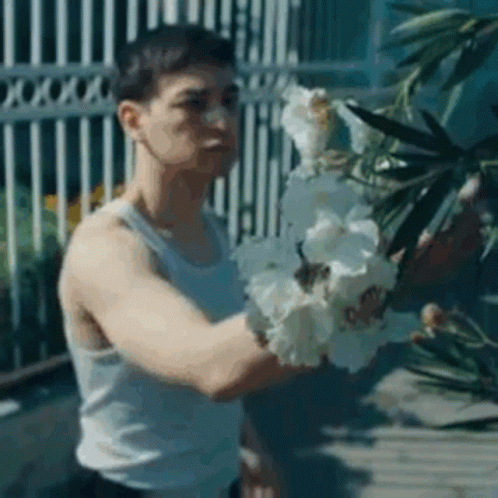 A Man In A White Tank Top Is Holding A Bunch Of White Flowers In His Hands . GIF