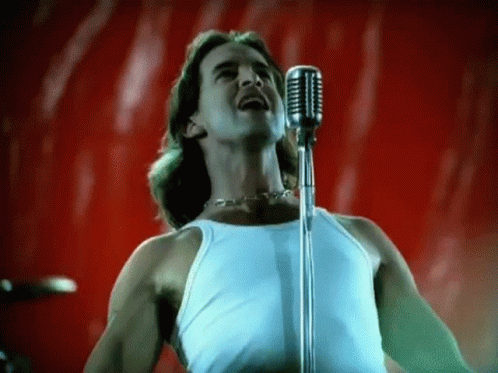 A Man In A White Tank Top Is Singing Into A Microphone On A Red Background . GIF