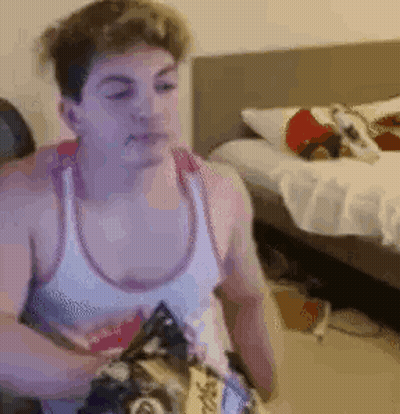 A Man In A White Tank Top Is Sitting In Front Of A Bed Eating Chips . GIF