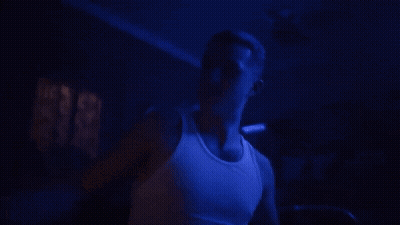 A Man In A White Tank Top Is Standing In A Dark Room With Blue Lights Behind Him . GIF