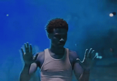 A Man In A White Tank Top Is Standing In A Dark Room With His Hands Up In The Air . GIF