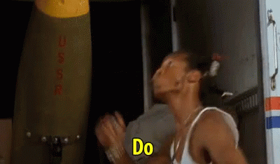 A Man In A White Tank Top Is Standing In Front Of A Large Bomb And Saying `` Do '' . GIF