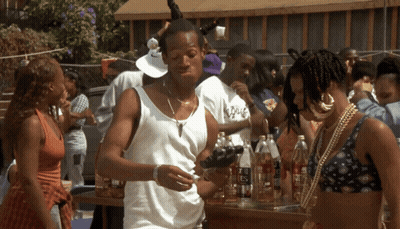 A Man In A White Tank Top Is Standing In Front Of A Table Full Of Bottles Including One That Says Es GIF