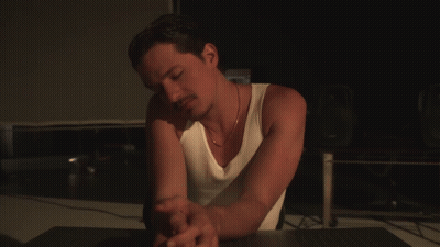 A Man In A White Tank Top Sits At A Desk GIF