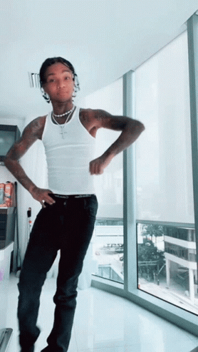 A Man In A White Tank Top Stands In Front Of A Large Window GIF