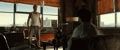 A Man In A White Tank Top Stands In Front Of A Large Window GIF