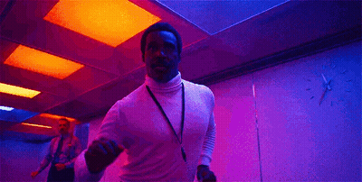 A Man In A White Turtleneck Is Dancing In A Pink Room With A Lanyard Around His Neck . GIF