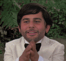 A Man In A White Tuxedo And Bow Tie Is Smiling With His Hands Folded GIF