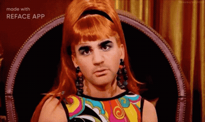 A Man In A Wig And Earrings Is Sitting In A Chair With A Colorful Dress On . GIF