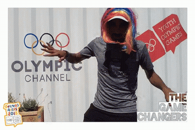 A Man In A Wig Is Dancing In Front Of A Sign For The Olympic Channel GIF