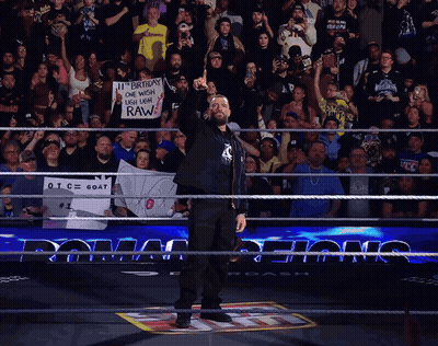 A Man In A Wrestling Ring Holds Up His Fist In Front Of A Crowd Holding A Sign That Says Raw GIF