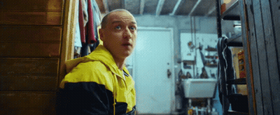 A Man In A Yellow And Black Jacket Is Standing In A Room Looking At Something . GIF
