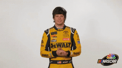 A Man In A Yellow And Black Racing Suit Is Standing In Front Of A Sign That Says What ? GIF