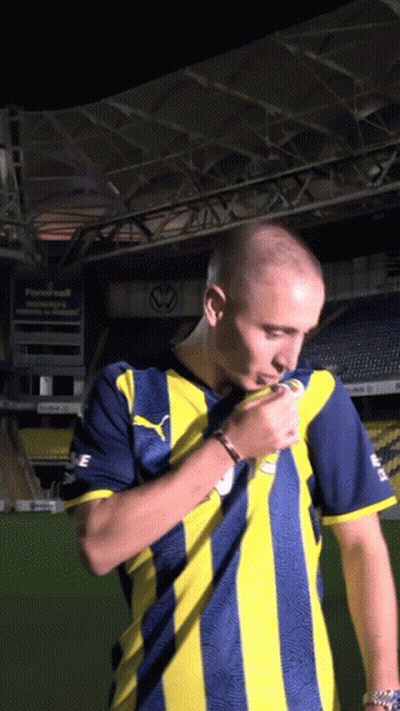 A Man In A Yellow And Blue Striped Shirt Stands In Front Of A Stadium GIF