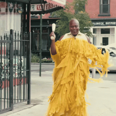 A Man In A Yellow Dress Is Walking Down The Street In Front Of A Store Called Ttleneck Outpost GIF