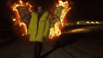 A Man In A Yellow Fur Coat Is Standing In Front Of Flames . GIF