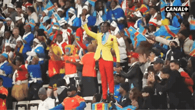A Man In A Yellow Jacket And Red Pants Stands In Front Of A Crowd With Canal + Written On The Bottom Right Corner GIF