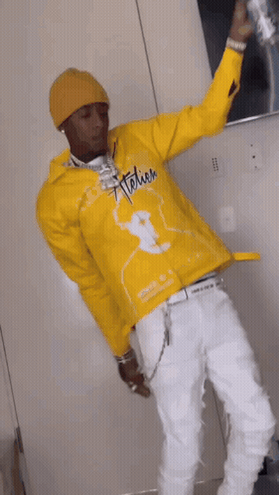 A Man In A Yellow Jacket And White Pants Is Standing In A Room Holding A Can Of Soda . GIF