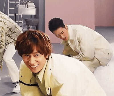 A Man In A Yellow Jacket Is Smiling While Another Man Squats Behind Him GIF