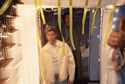 A Man In A Yellow Jacket Is Standing In A Hallway With Ribbons Hanging From The Ceiling GIF