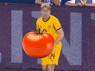 A Man In A Yellow Jersey Is Holding A Large Tomato In Front Of A Sign That Says Ucl GIF