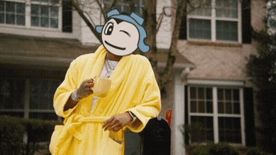 A Man In A Yellow Robe Holds A Cup Of Coffee In Front Of A House GIF