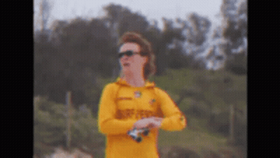 A Man In A Yellow Shirt And Sunglasses Is Standing On A Beach Holding Binoculars . GIF