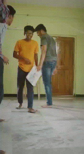 A Man In A Yellow Shirt Is Laying On The Floor While Another Man Stands In The Background . GIF