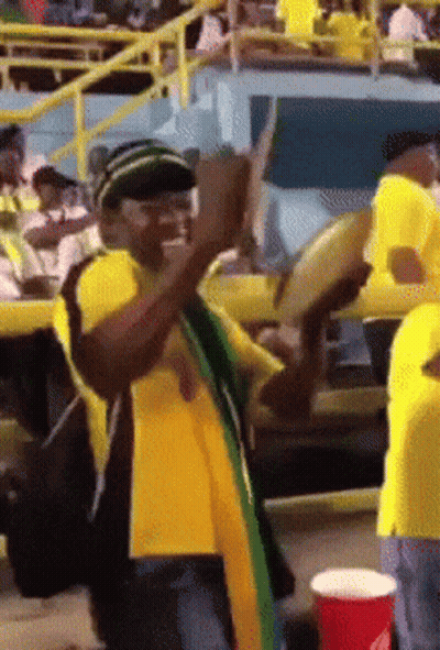 A Man In A Yellow Shirt Is Playing A Drum In A Crowd GIF