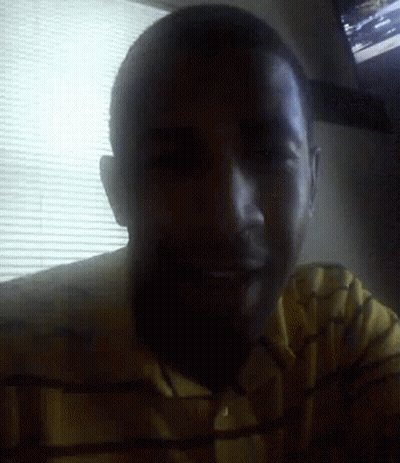 A Man In A Yellow Shirt Is Smiling In Front Of A Window With Blinds GIF
