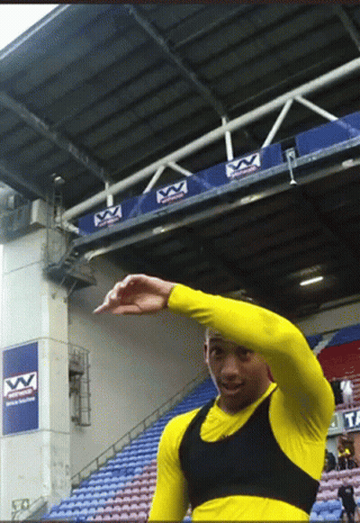A Man In A Yellow Shirt Is Standing In Front Of A Stadium With A Sign That Says Vv On It GIF