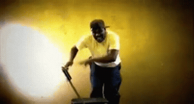A Man In A Yellow Shirt Is Standing Next To A Suitcase And A Mop . GIF