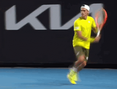 A Man In A Yellow Shirt Is Swinging A Tennis Racket In Front Of A Kia Logo GIF