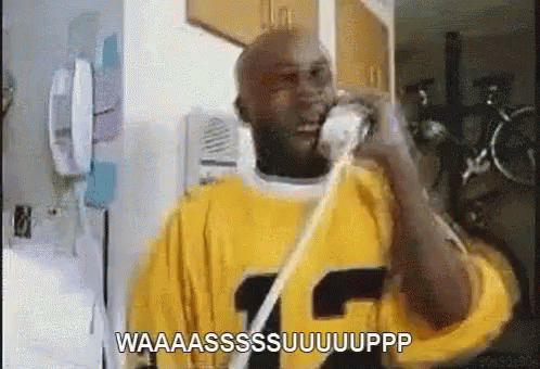 A Man In A Yellow Shirt Is Talking On A Phone And Says Waaaassssuuuuupp GIF