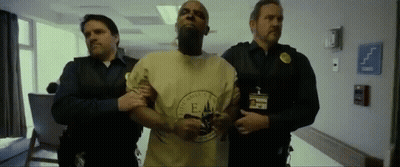 A Man In A Yellow Shirt With The Letter E On It Is Being Led By Two Men GIF